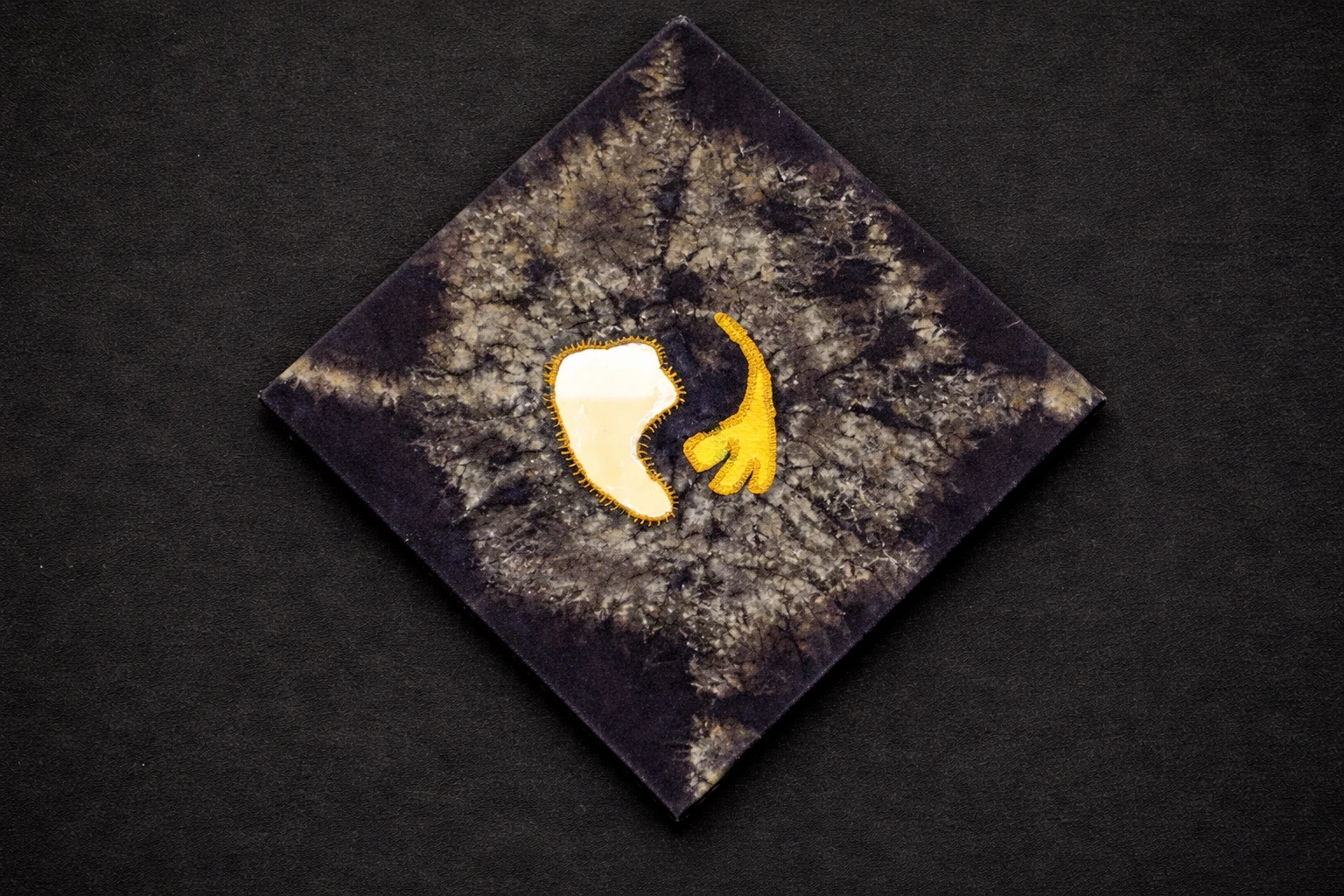 A diamond-shaped textile with a small mirror and yellow stitched hand